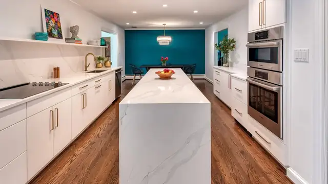 Galley kitchen with a white waterfall quartz island, flat white cabinets and a teal accent wall